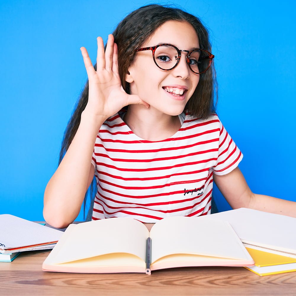 Young girl student sitting at desk with notebook in front of her holding her hand up to her ear