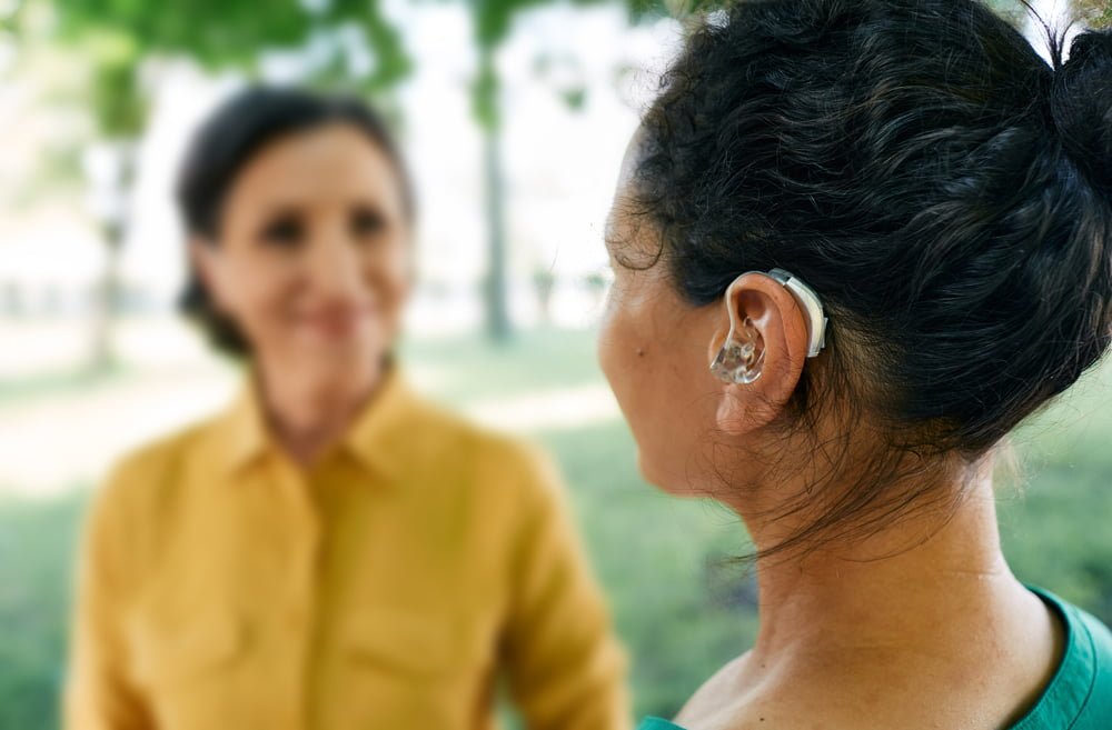 Adult woman with hearing loss uses a hearing aid to communicate with her female friend at city park